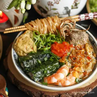 A bowl of Hai Phong banh da cua with broad red rice noodles, greens and assorted toppings