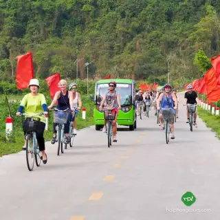 Cyclists and an electric cart on a paved road in the Viet Hai area of Cat Ba